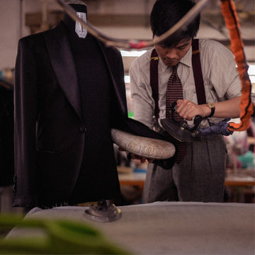 A tailor pressing the sleeve of a dark tailored jacket on a dress form using a steam iron, with sewing workshop tools and fabric visible in the background.