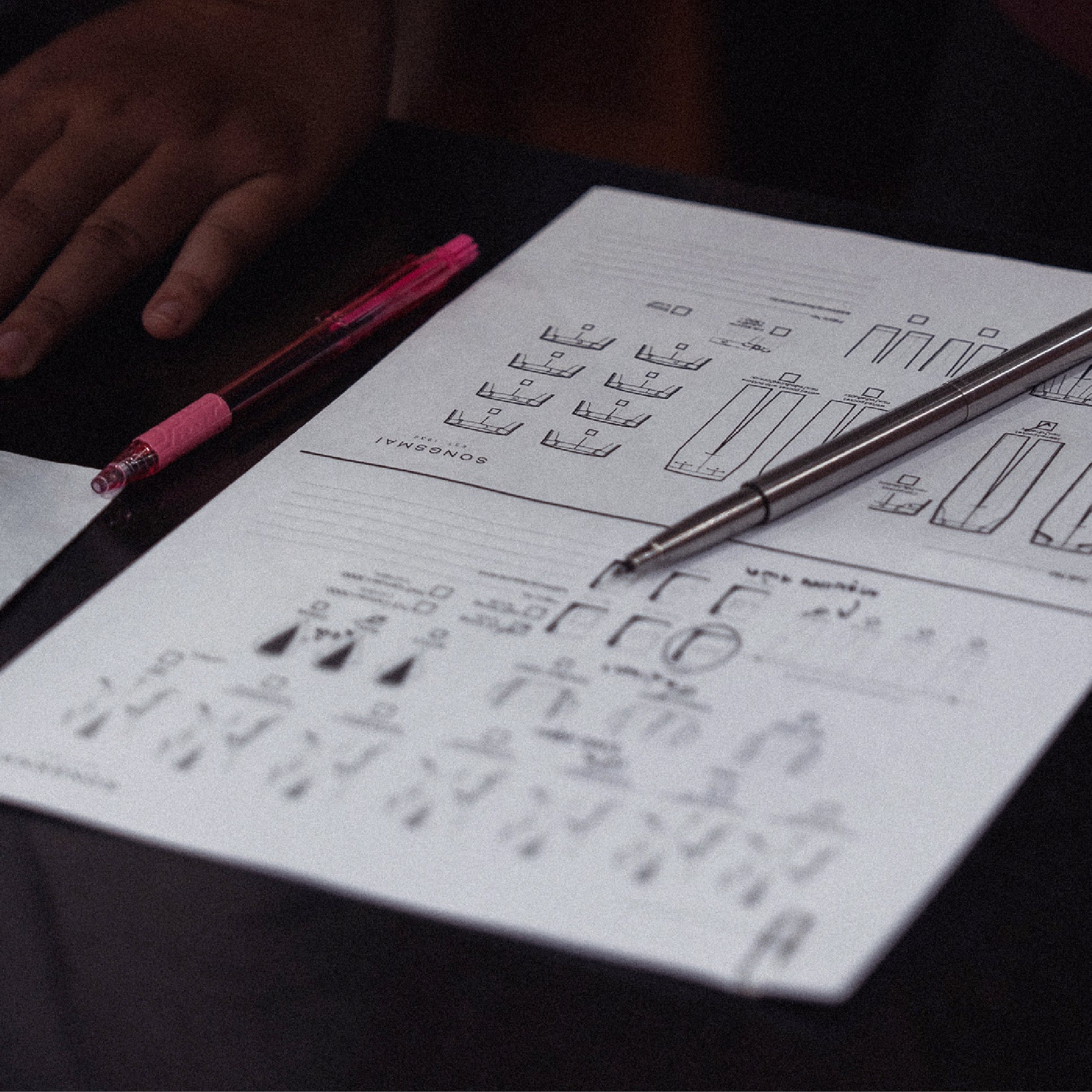 A close-up of bespoke suit sketches and measurement diagrams on paper, with a silver pen and a pink mechanical pencil resting on a dark tabletop.
