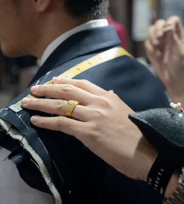 A tailor measuring a client’s shoulder with a yellow measuring tape while holding the jacket canvas in place; the image highlights the tailor’s hand with a gold ring, pins on a wrist pincushion, and the precision of shoulder fitting during a bespoke tailoring fitting.