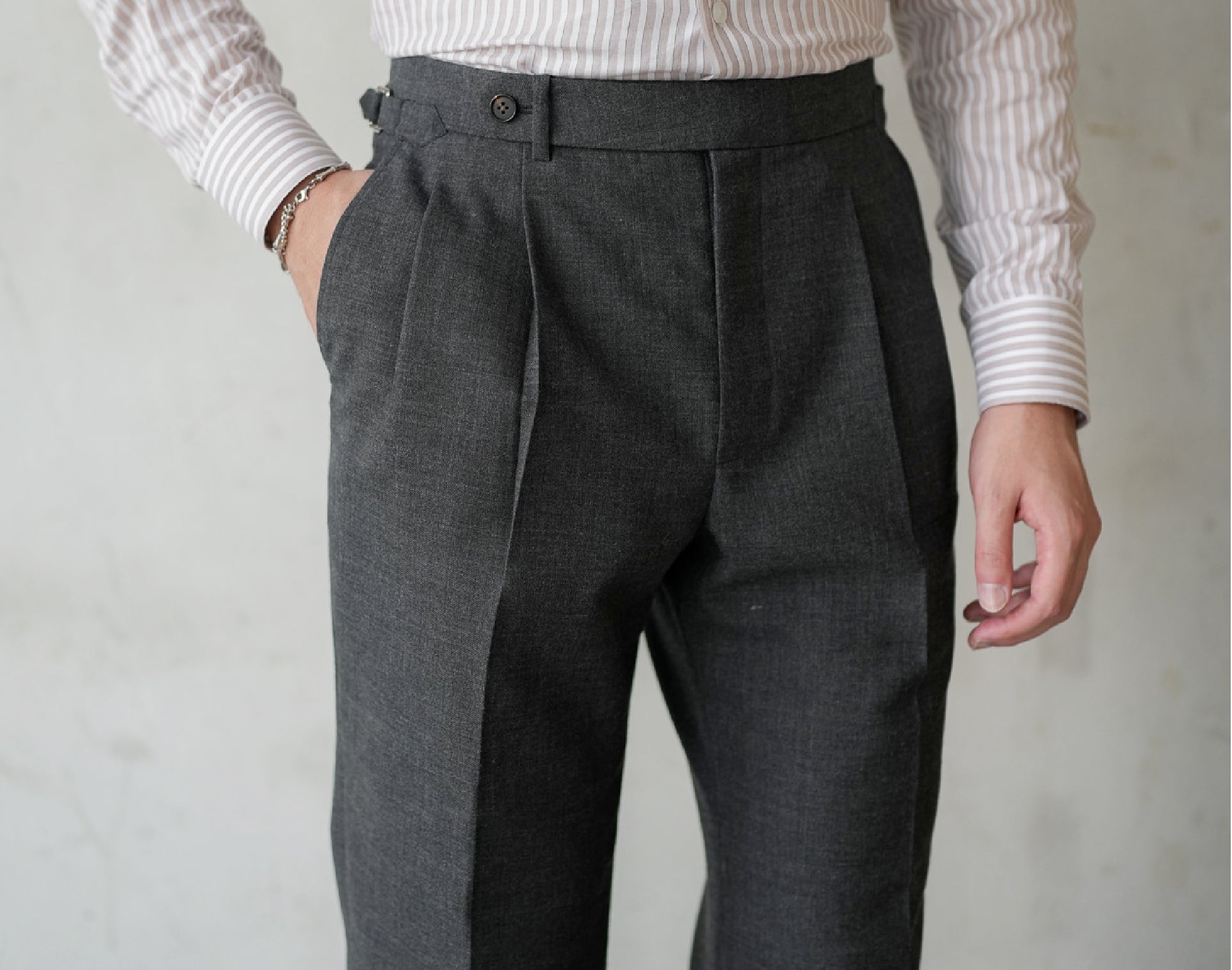 A close-up of a man wearing dark grey pleated tailored trousers with side adjuster tabs, a tucked striped shirt, and one hand in the pocket—highlighting the clean front crease, fabric texture, and refined tailoring details.