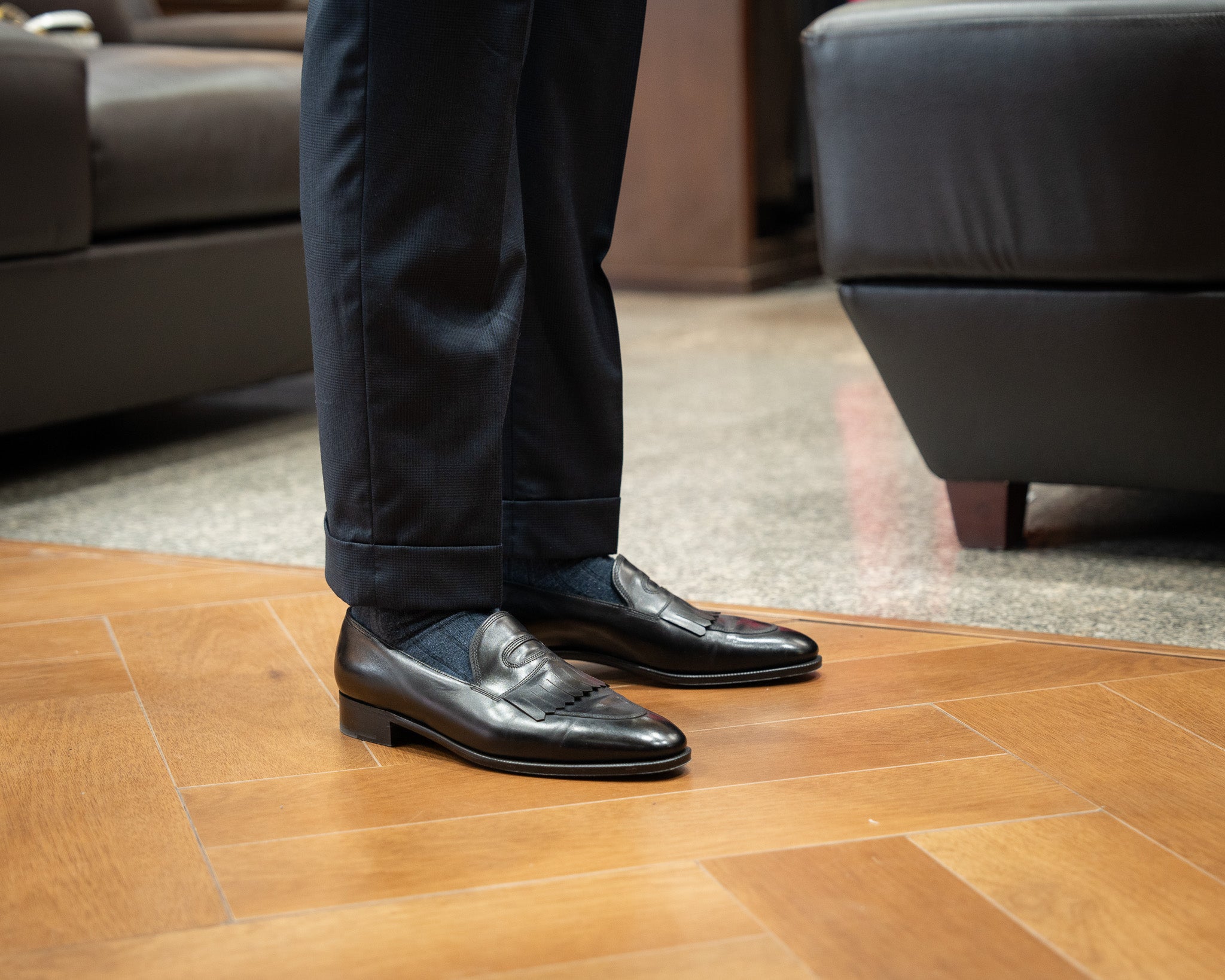 Person wearing black dress shoes and dark pants standing on a wooden floor.