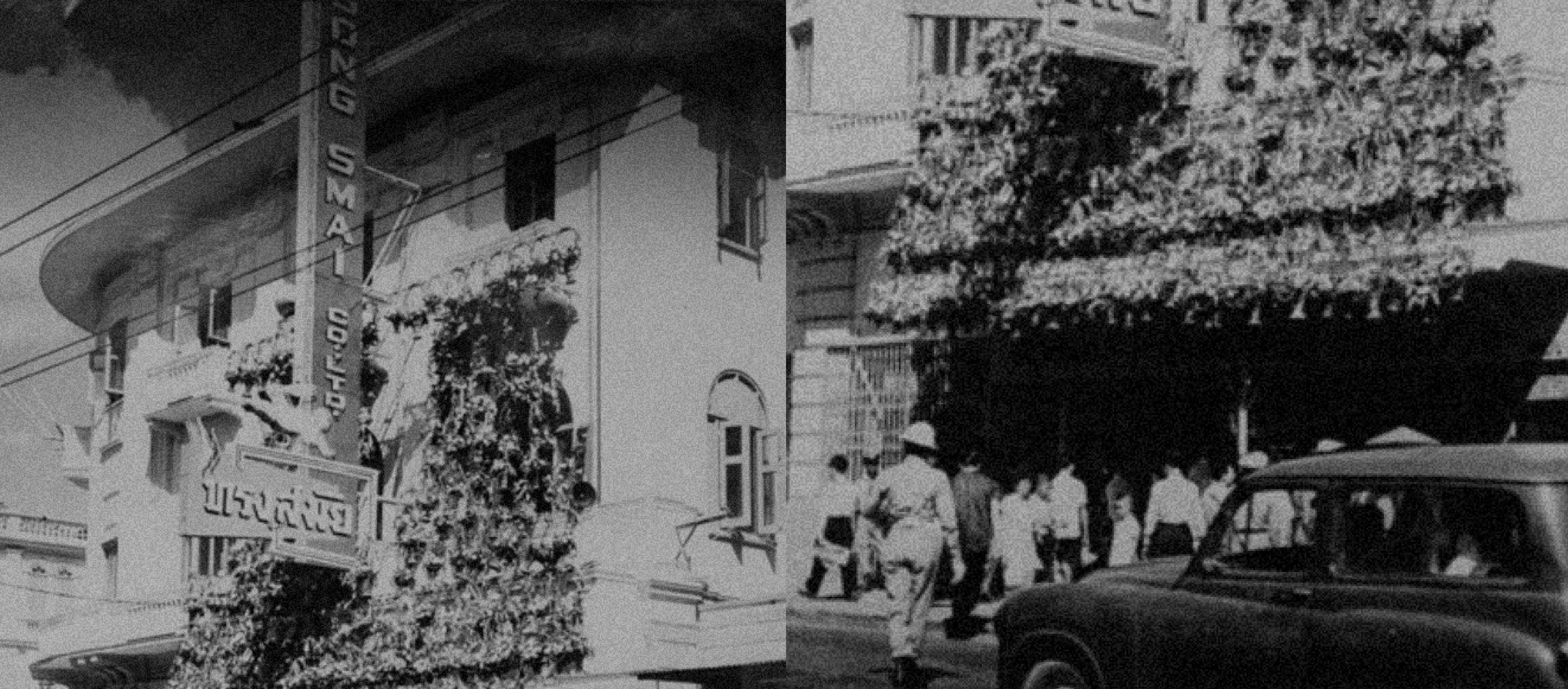 A vintage black-and-white street scene showing the exterior of an old Songsmai tailoring shop with vertical signage, decorative awnings, pedestrians walking along the sidewalk, and a parked classic car in the foreground — evoking the brand’s long history and storefront presence.